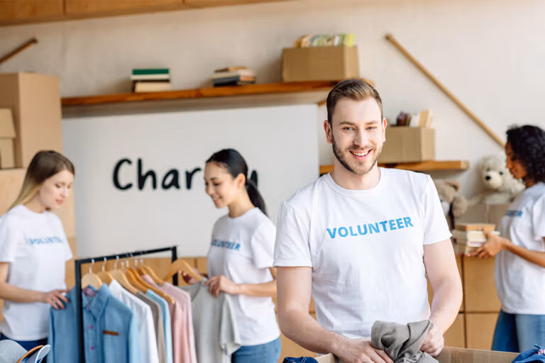 Group of people sorting clothing indoors – smiling man in foreground holding folded garment.