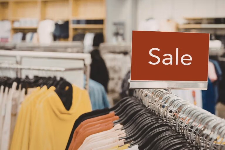 A clothes shop showing different coloured jumpers on a rail with a red "Sale" sign ono top.
